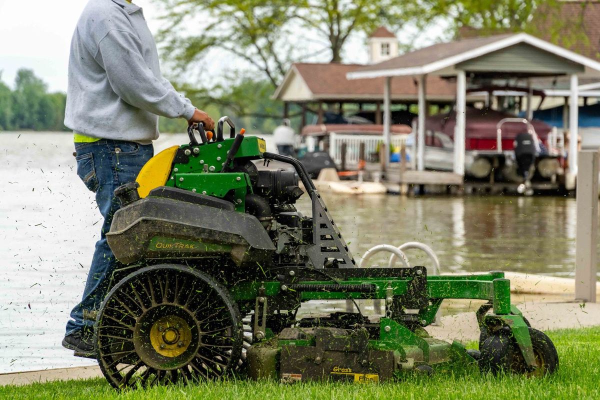 John Deere QuikTrak stand-on mower cutting a lakefront lawn