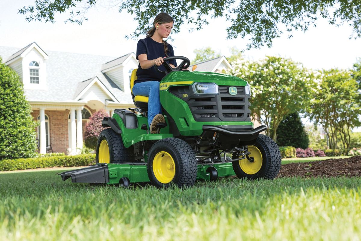 Woman driving a John Deere S160 lawn tractor on a green lawn in front of a house