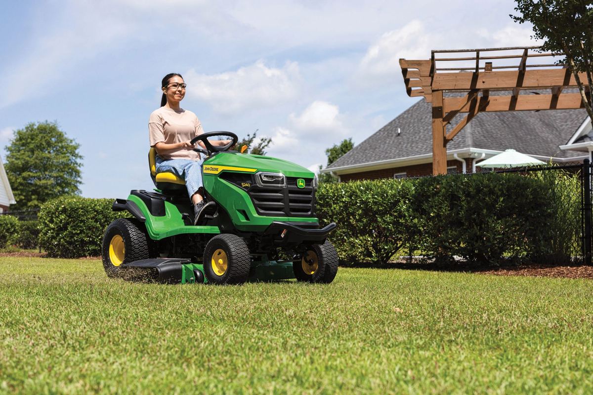 Woman in glasses driving a John Deere S140 riding lawn mower on a green lawn with a house in the background