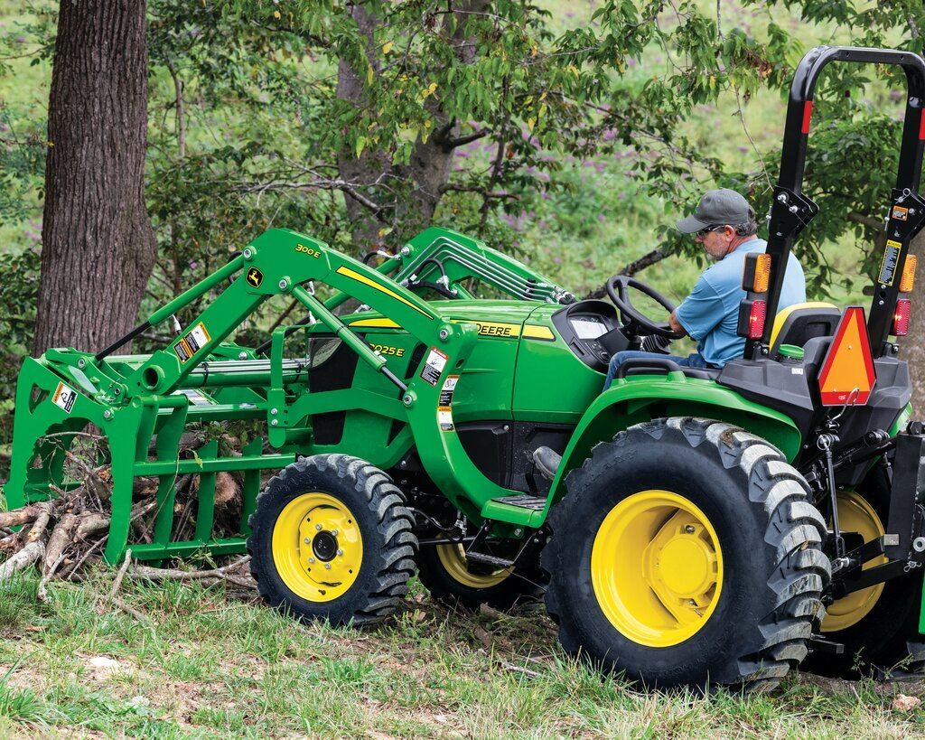 Man operating a John Deere 3025E compact tractor with a front loader and grapple attachment moving brush