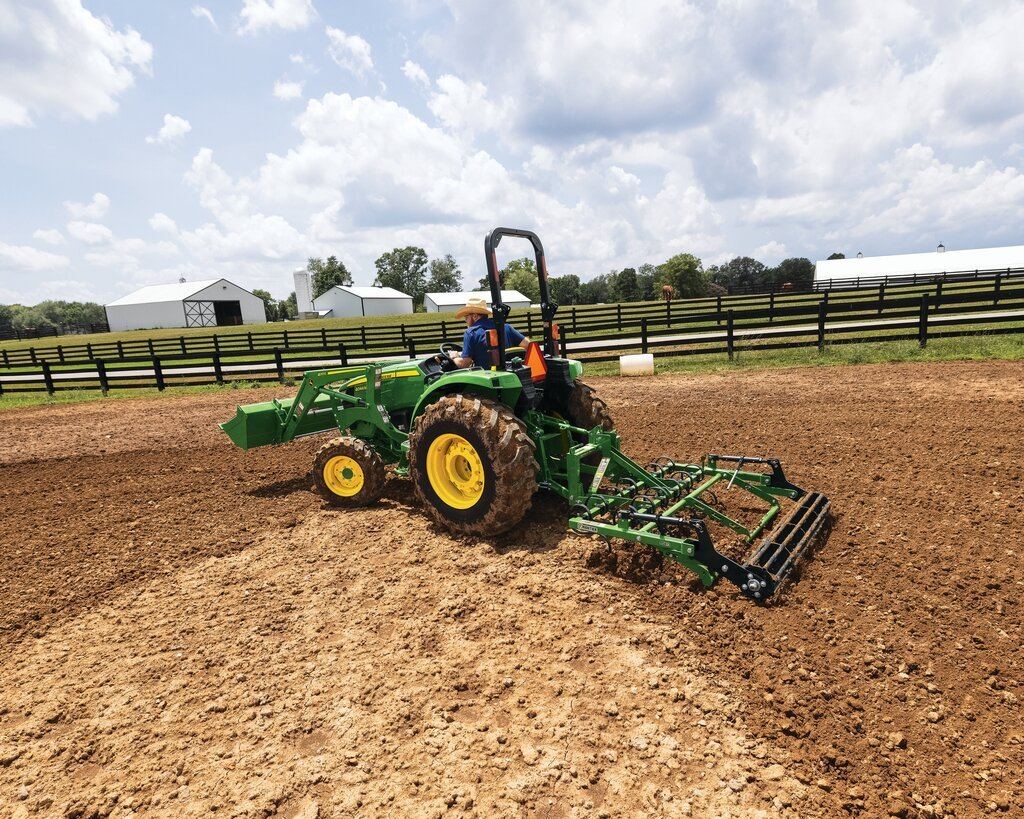 Man on a green John Deere tractor with front loader and rear implement tilling a brown field near white barns
