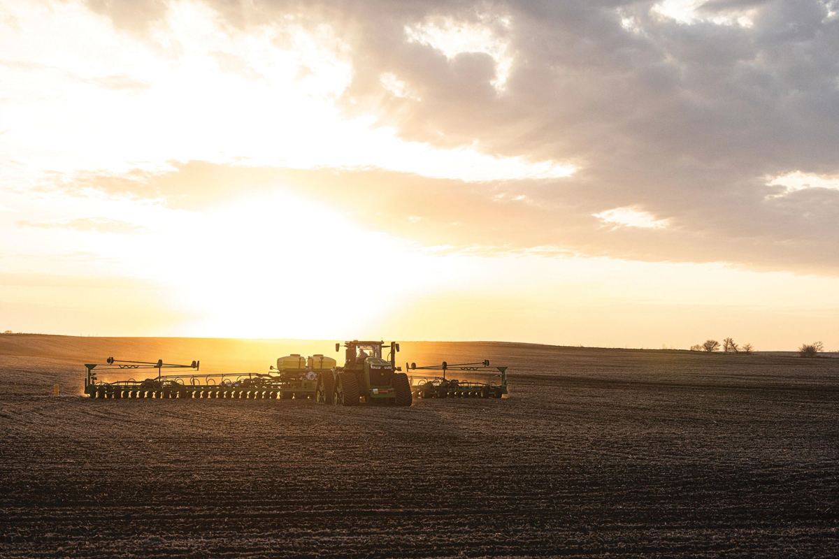 A John Deere 9R tractor is shown pulling a large air seeder across a vast field under a dramatic sunset sky