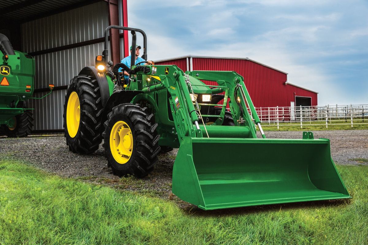 Man driving a green John Deere tractor with H250 front loader and yellow wheels, next to another green machine and a red barn