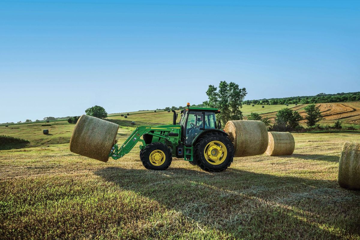 Green John Deere 6R tractor with front loader carrying a hay bale in a field with more bales
