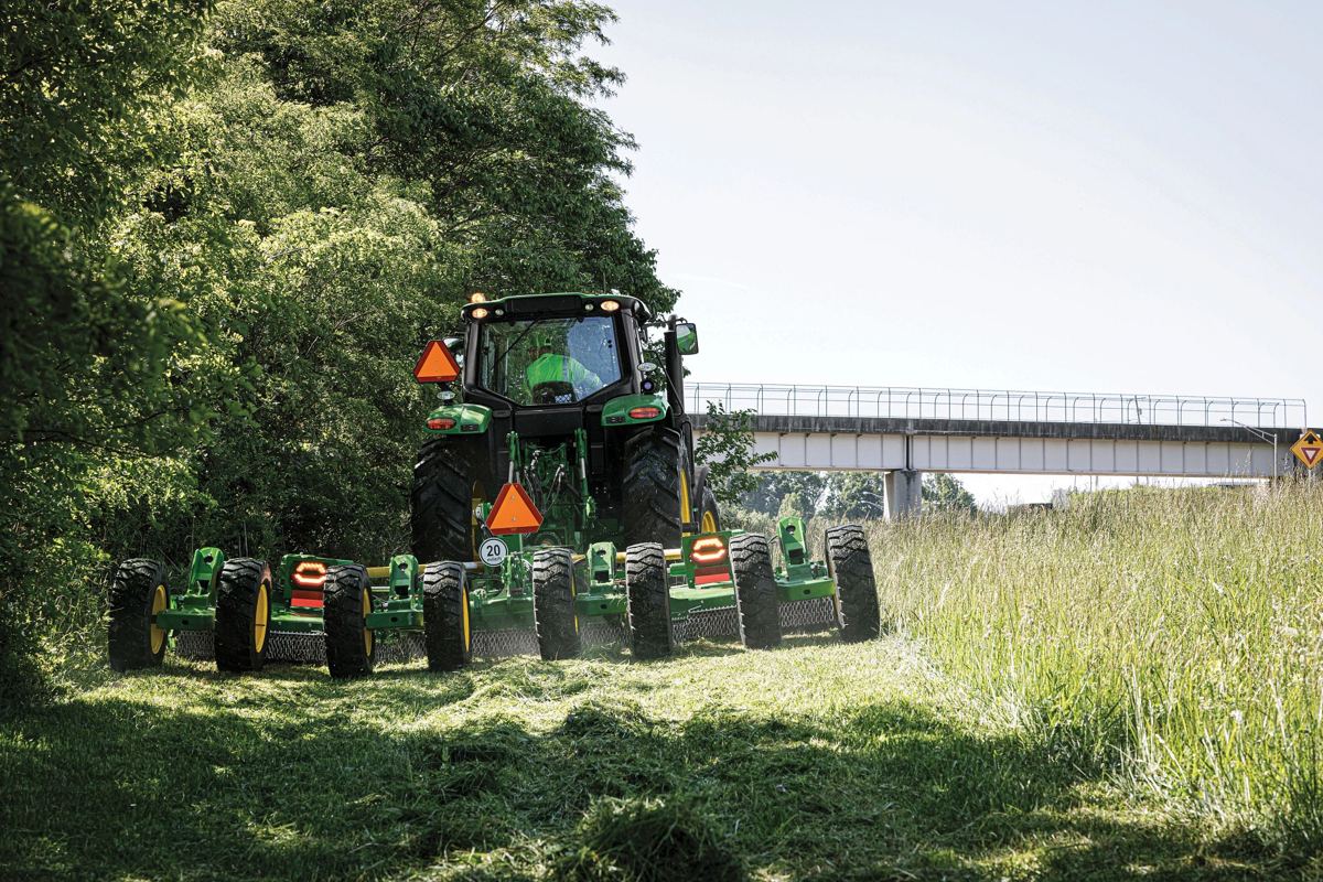 A John Deere 6M tractor is actively mowing a field with a multi-unit mower conditioner, fitting the description of work on a hay farm in a Midwest setting