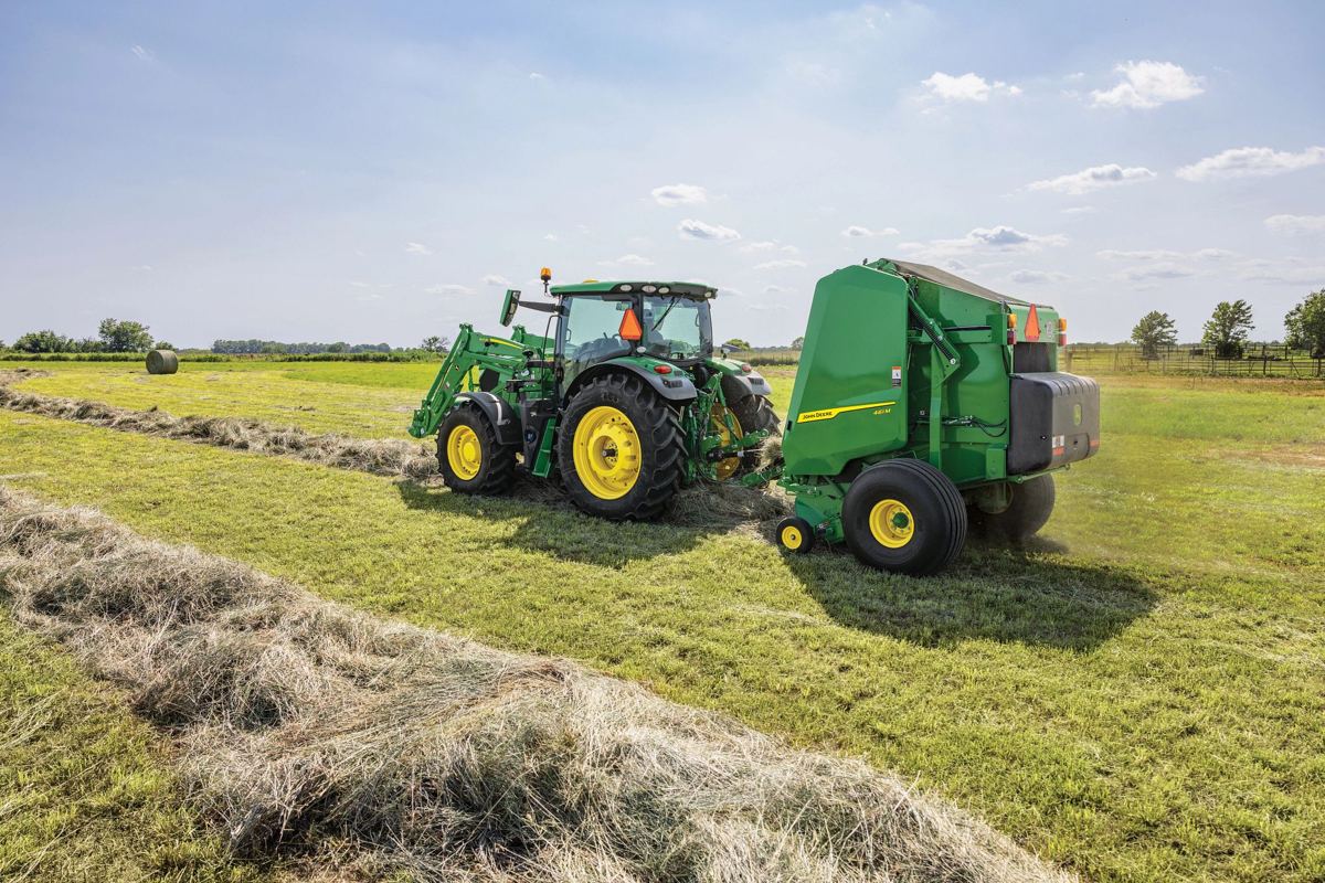 A John Deere 6R series tractor is shown actively baling hay in a field under a clear sky