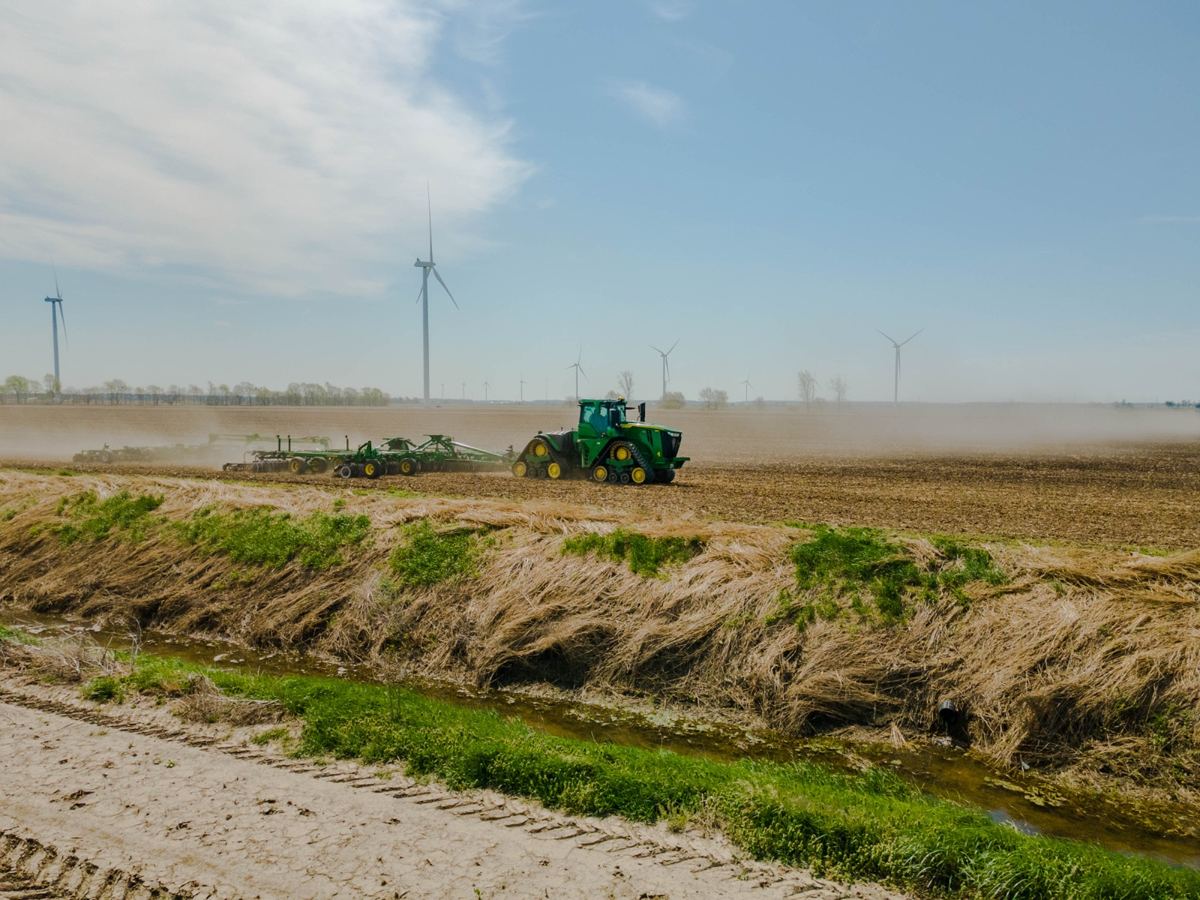 John Deere tractor tilling a field with a planter, dust rising, wind turbines in the background
