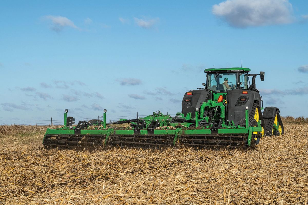 A John Deere 8RX four-track tractor is pulling a large tillage tool through a field of corn stubble under a clear blue sky