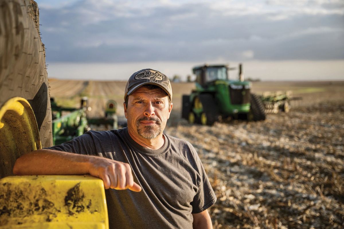 Man in a John Deere cap leaning on yellow farm machinery, with a green John Deere track tractor in a harvested field