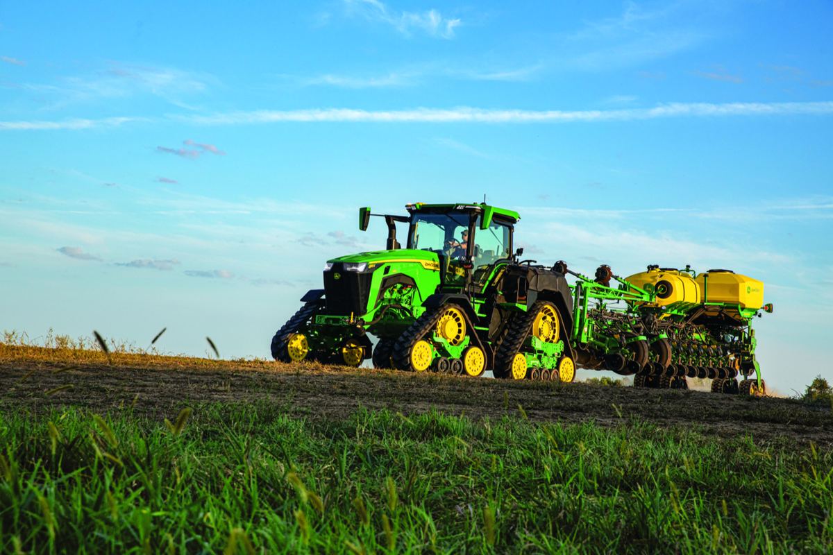 Person driving a John Deere 8RX track tractor pulling a planter in a field with green grass and blue sky