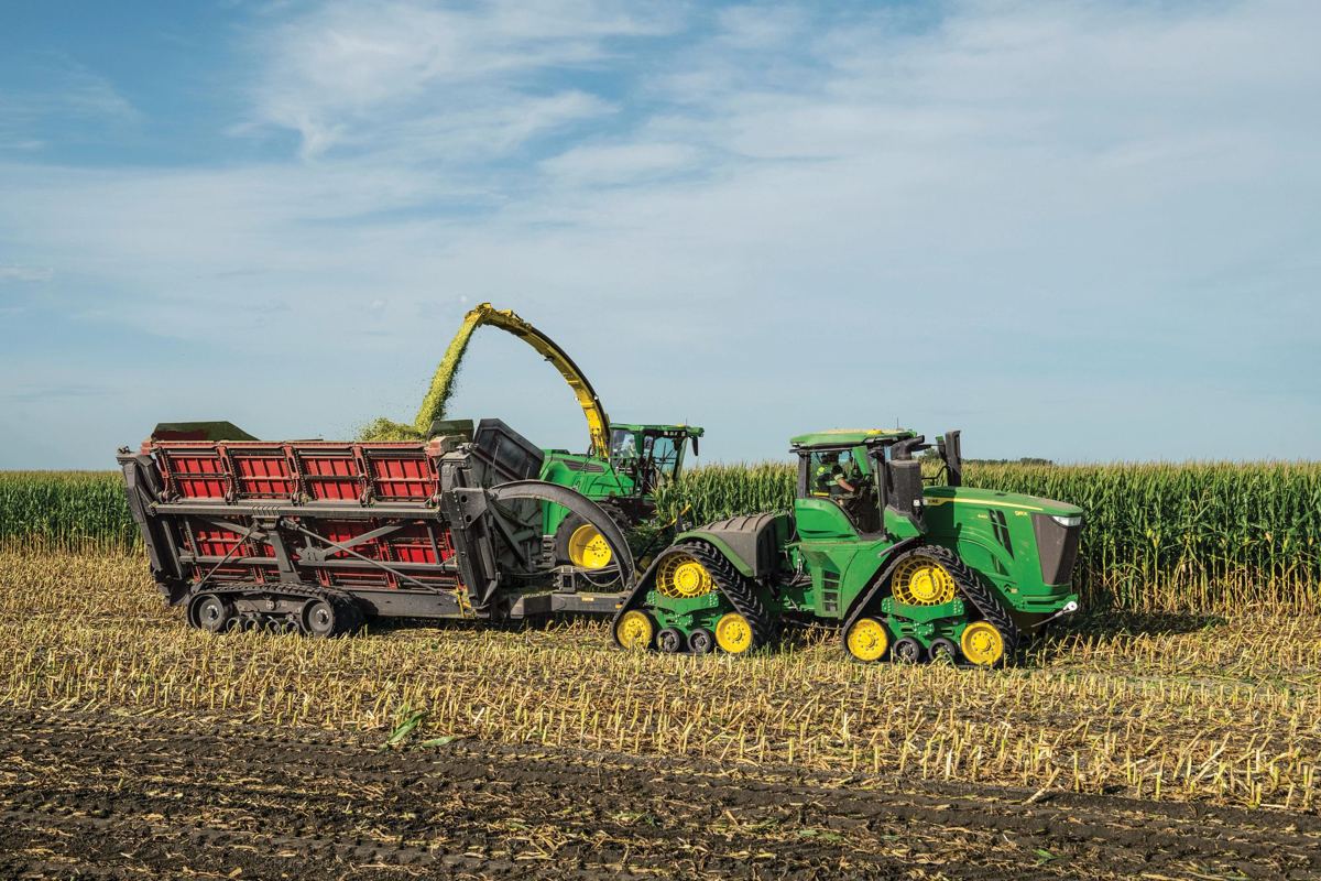 John Deere 9RX tractor pulling a forage wagon while a John Deere harvester fills it with chopped corn