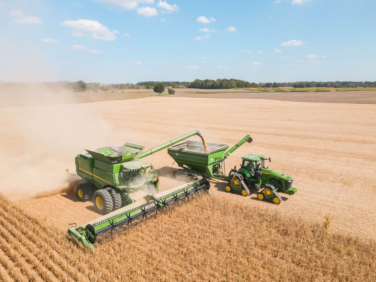 John Deere combine harvesting a field, offloading grain into a grain cart pulled by a John Deere tractor
