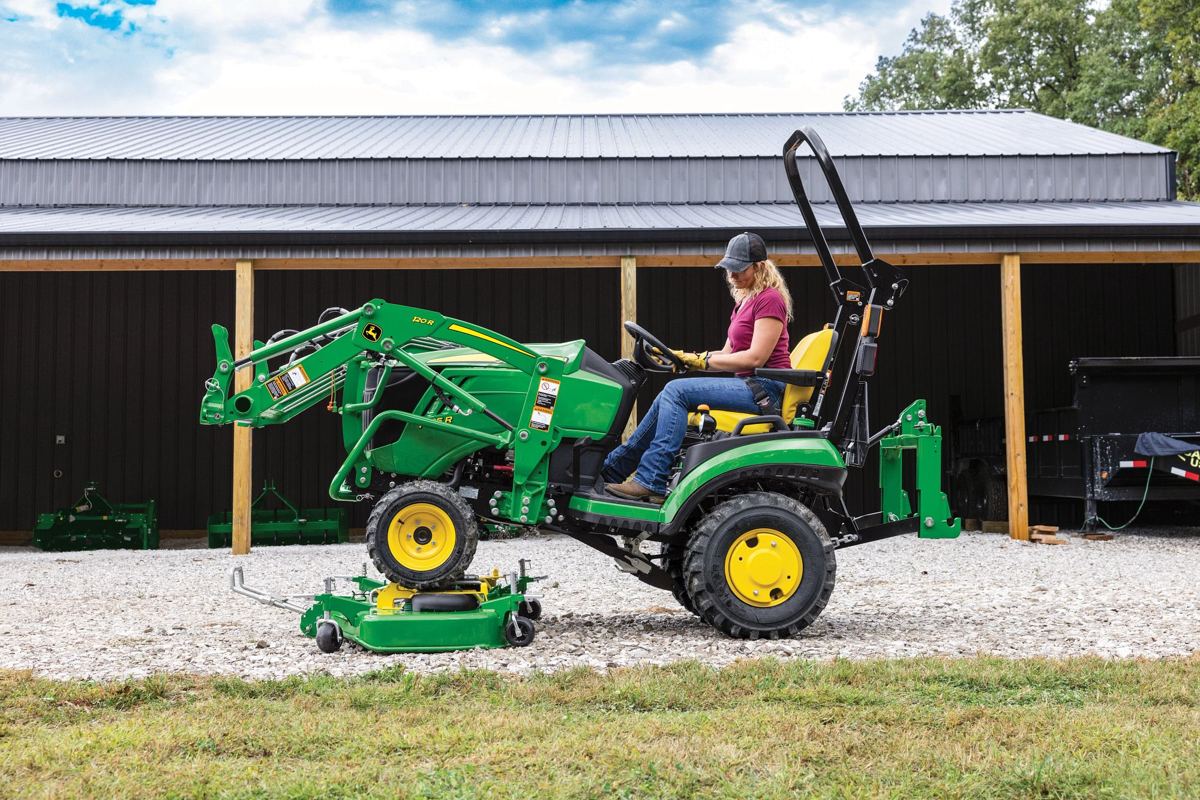 Woman operating a green John Deere tractor with a 120R front loader and mid-mount mower