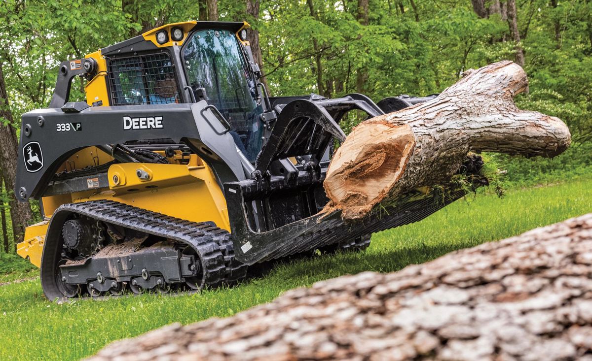 A John Deere compact track loader uses a grapple attachment to carry a large log up a grassy incline, demonstrating the equipment in action on the job