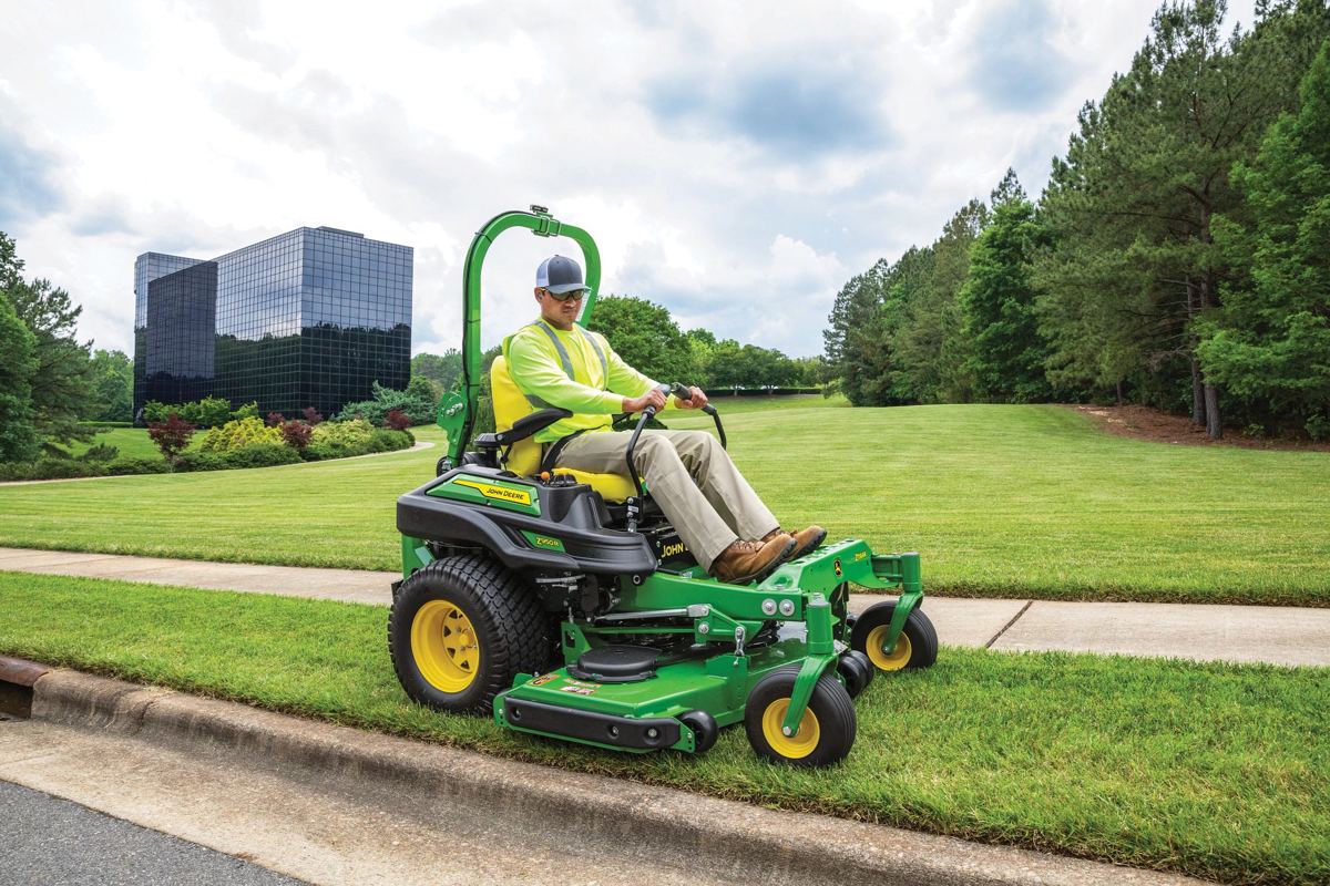 Man on a John Deere Z950M zero-turn mower cutting grass next to a curb with a building in the background