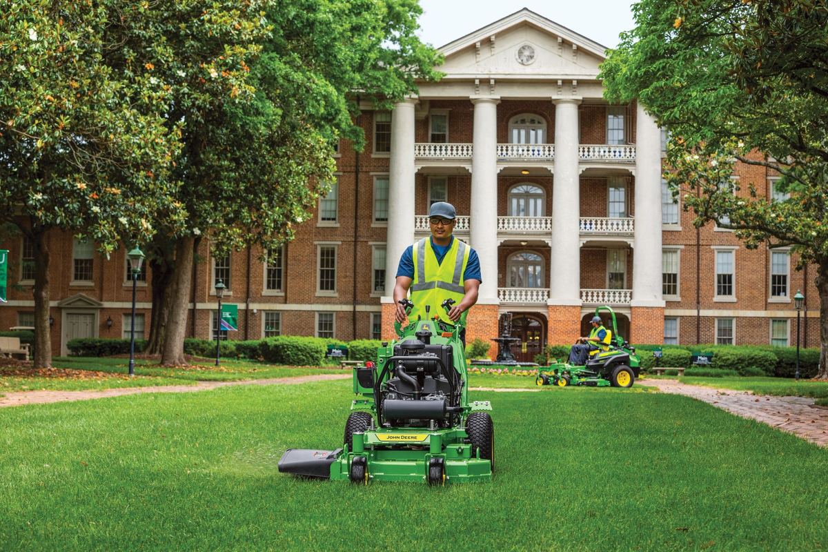 Two men in safety vests operate John Deere mowers on a green lawn in front of a large brick building