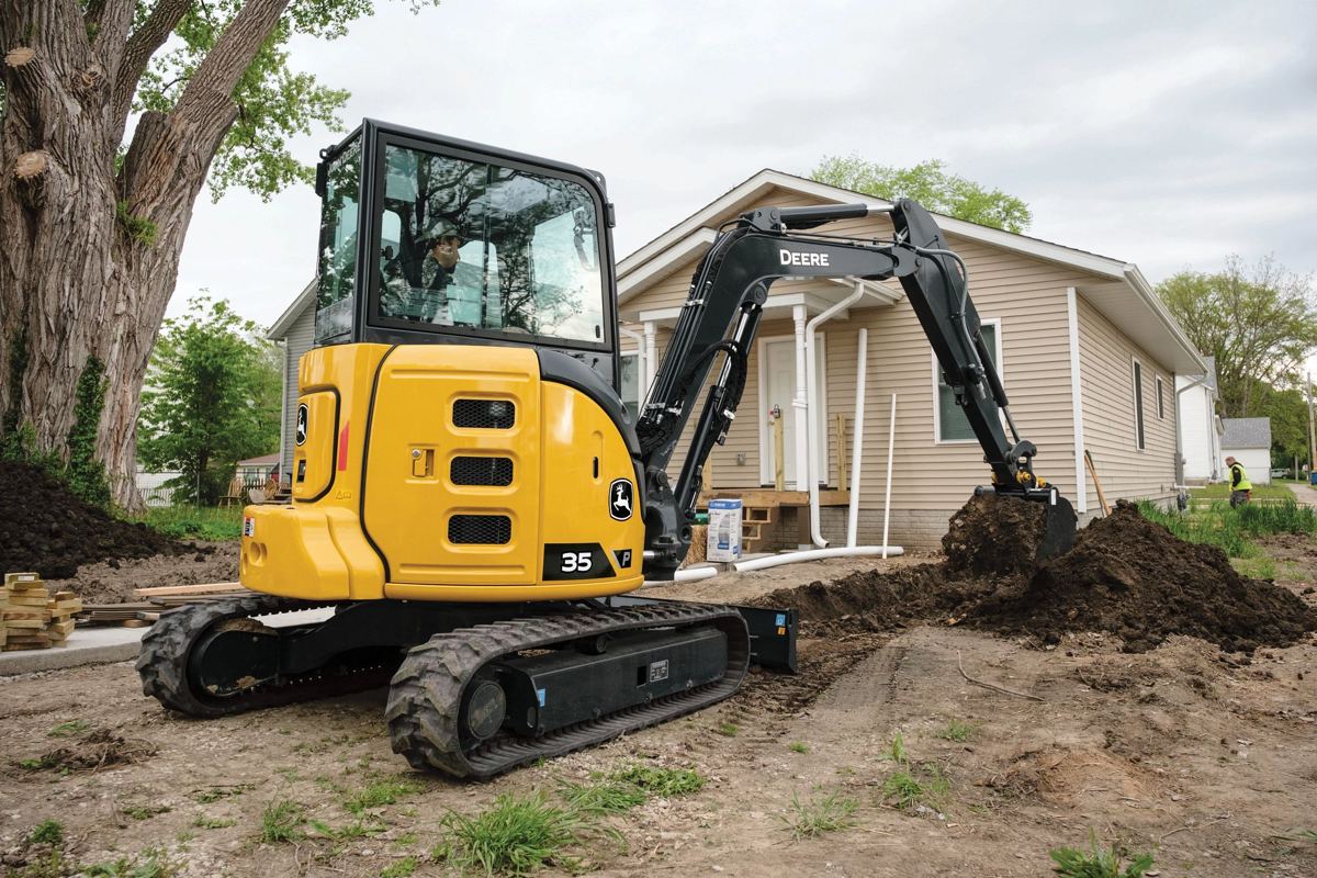 A John Deere 35 compact excavator is shown digging a trench next to a residential house, with its bucket actively in the ground