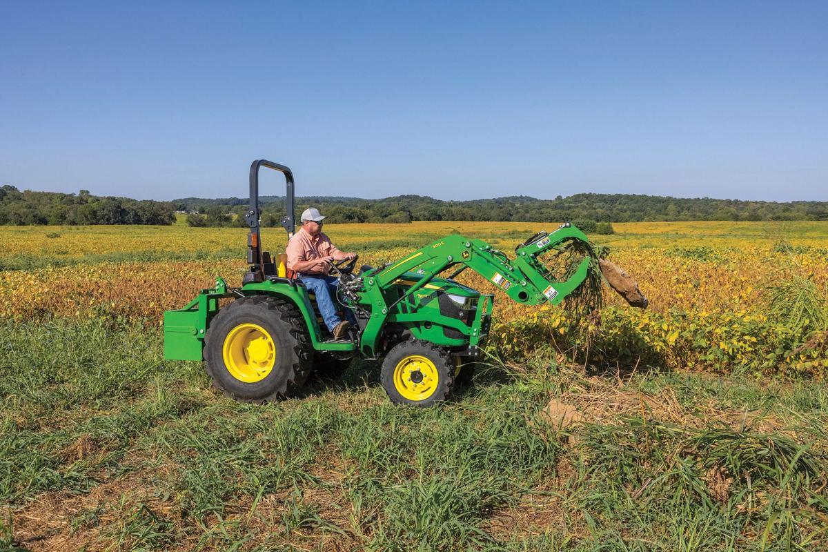 A John Deere compact tractor with a front loader is actively moving vegetation or debris in a field, without a backhoe attachment