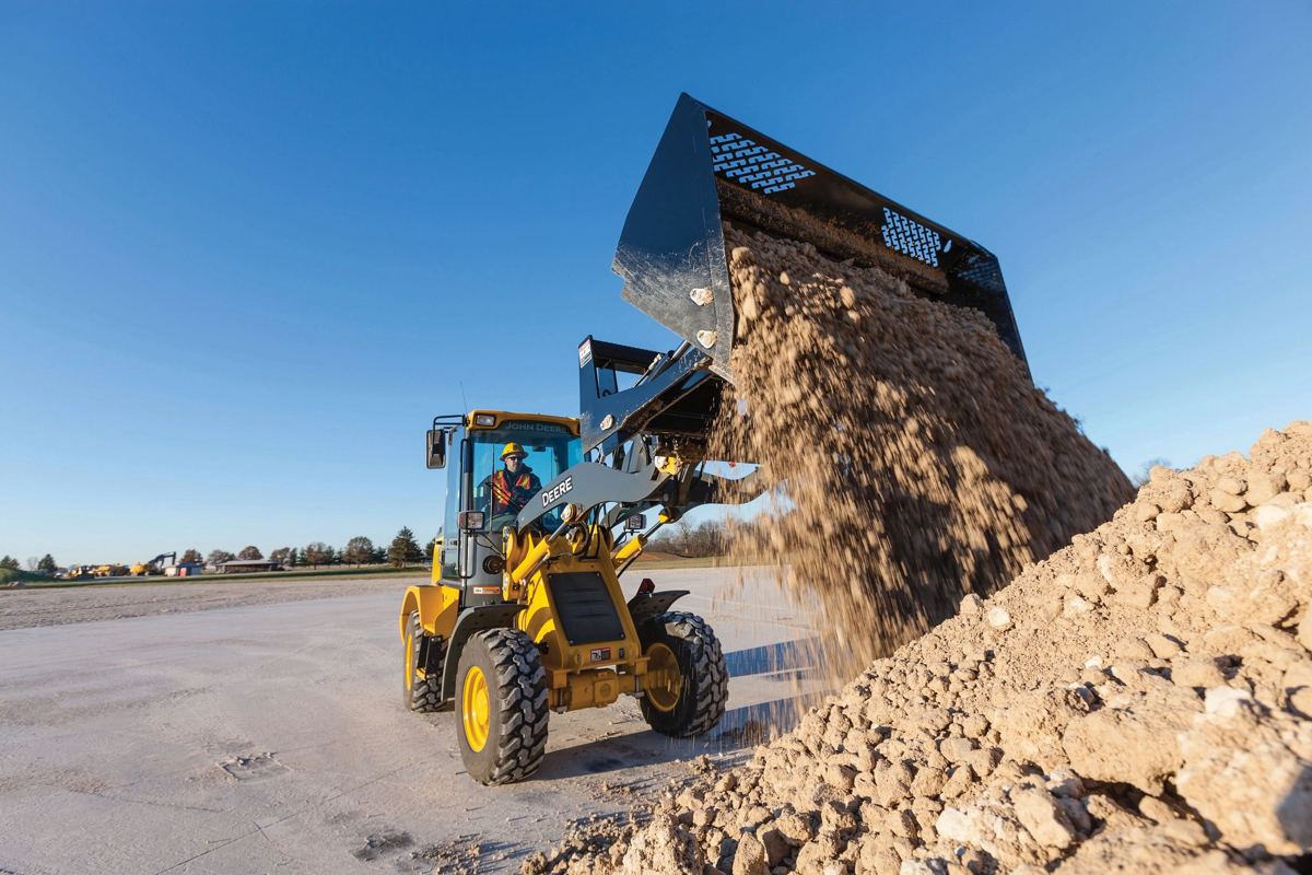 Yellow John Deere wheel loader operated by a person in a hard hat, dumping a large pile of dirt onto the ground