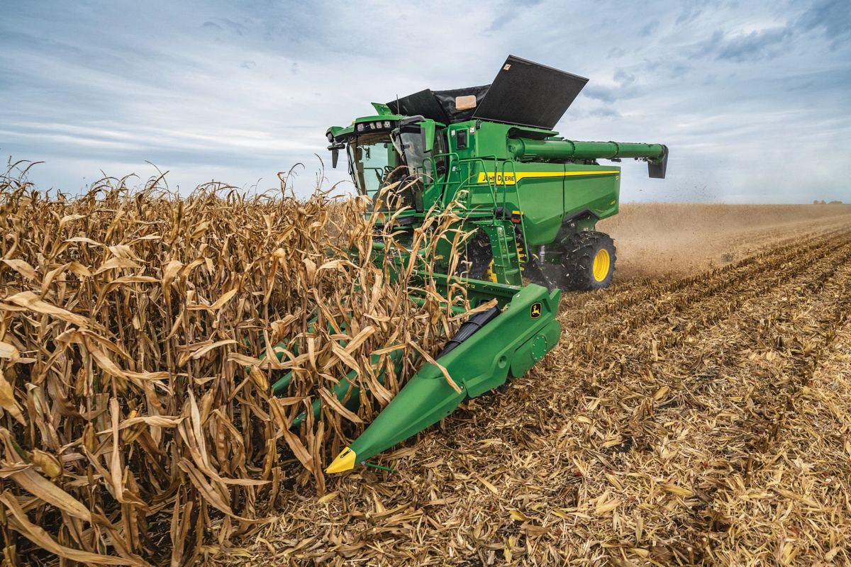 Green John Deere combine harvester with a corn head working in a field of dry corn under a cloudy sky