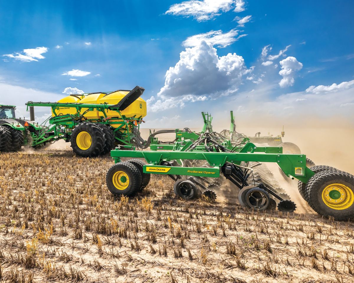 John Deere tractor pulling a yellow air cart and a green ProSeries Opener kicking up dust in a field