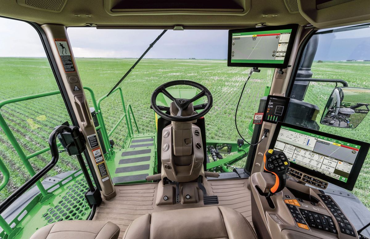 Interior view of a John Deere combine cab with steering wheel, multiple screens, and controls, looking out at a green field