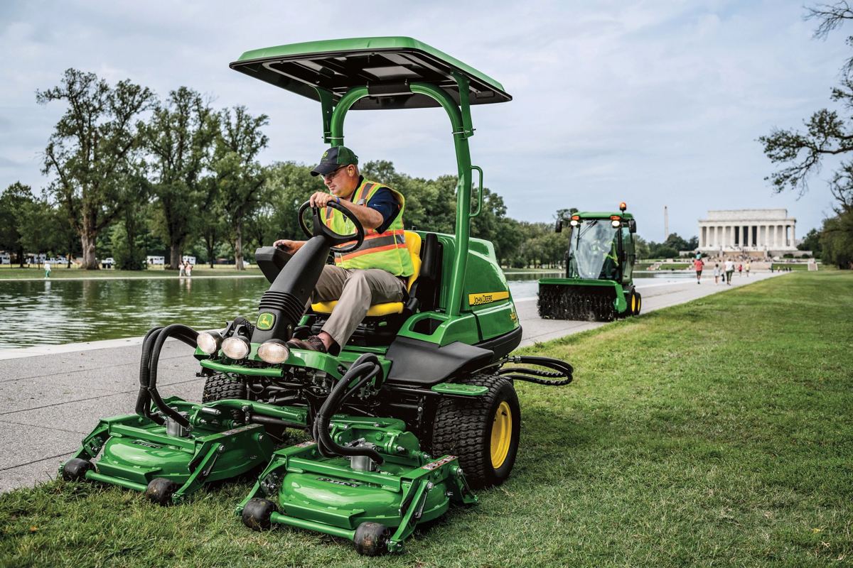 A John Deere 1500 Series TerrainCut front mower actively mowing grass along a path in a park setting with trees and the Lincoln Memorial in the background