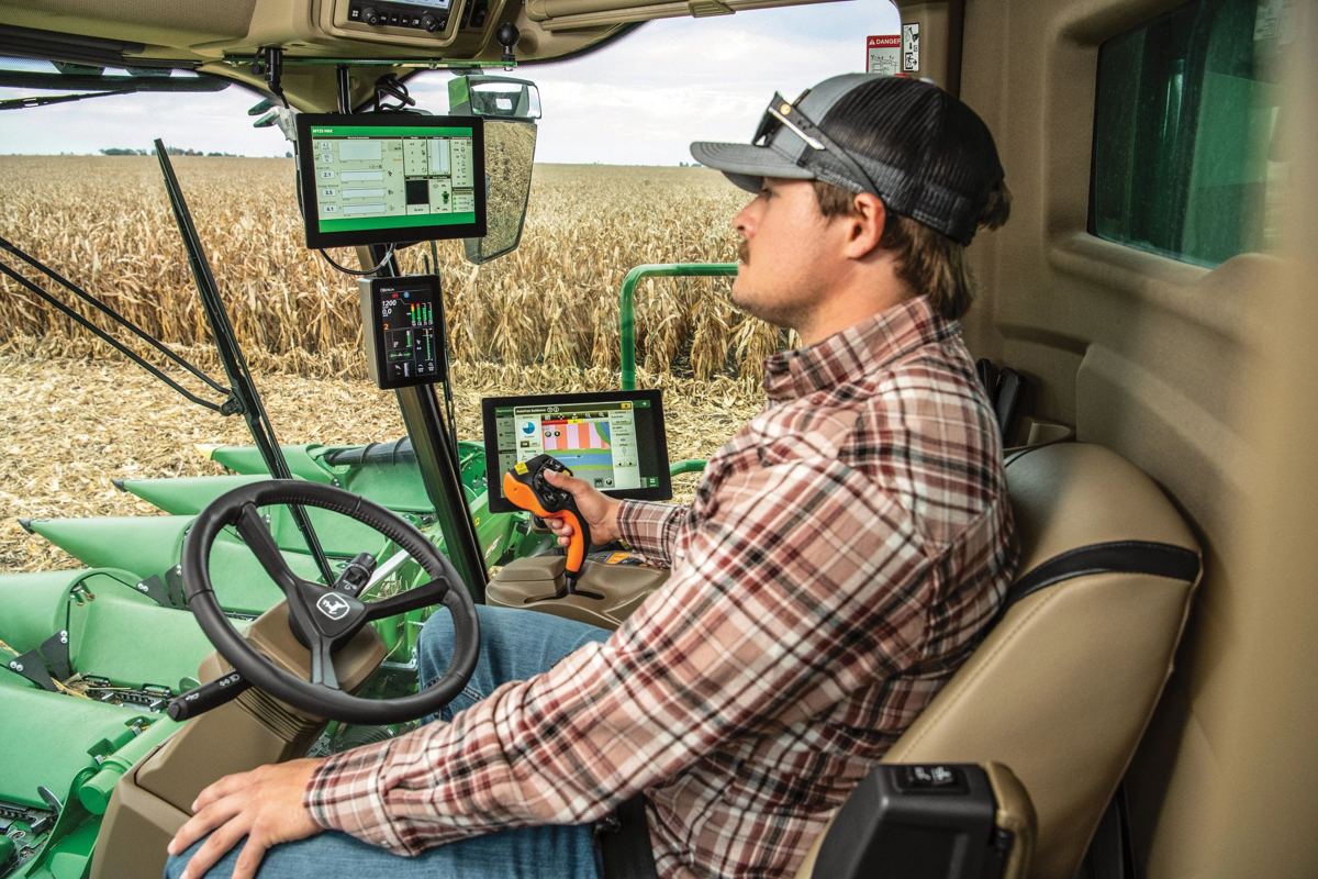 A farmer operates a modern John Deere combine in a cornfield, with the combine's control screens visible