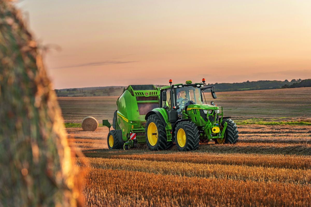 Green John Deere tractor pulling a baler in a harvested field with a person driving at sunset