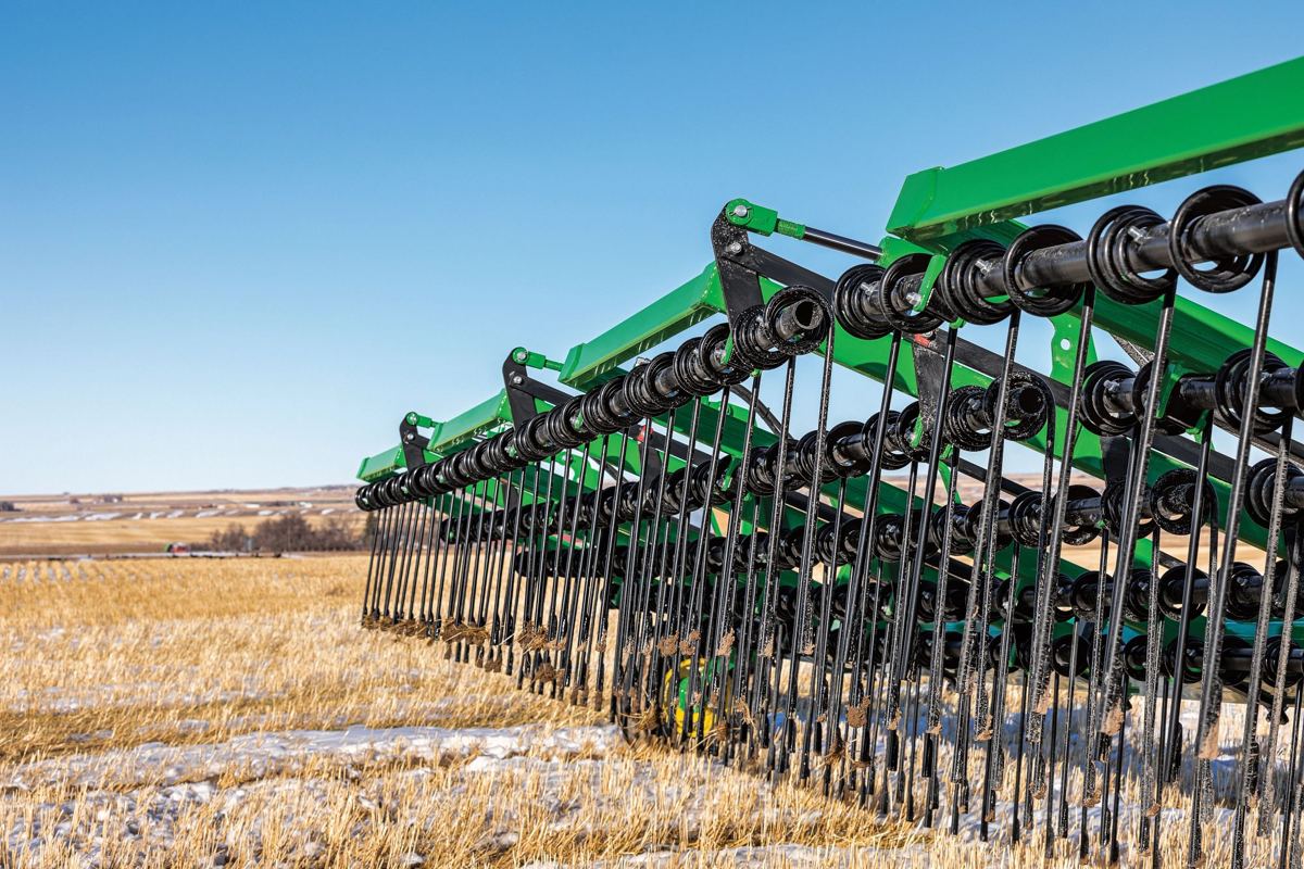 Close-up of a green agricultural harrow with black tines in a field of golden stubble and snow under a blue sky