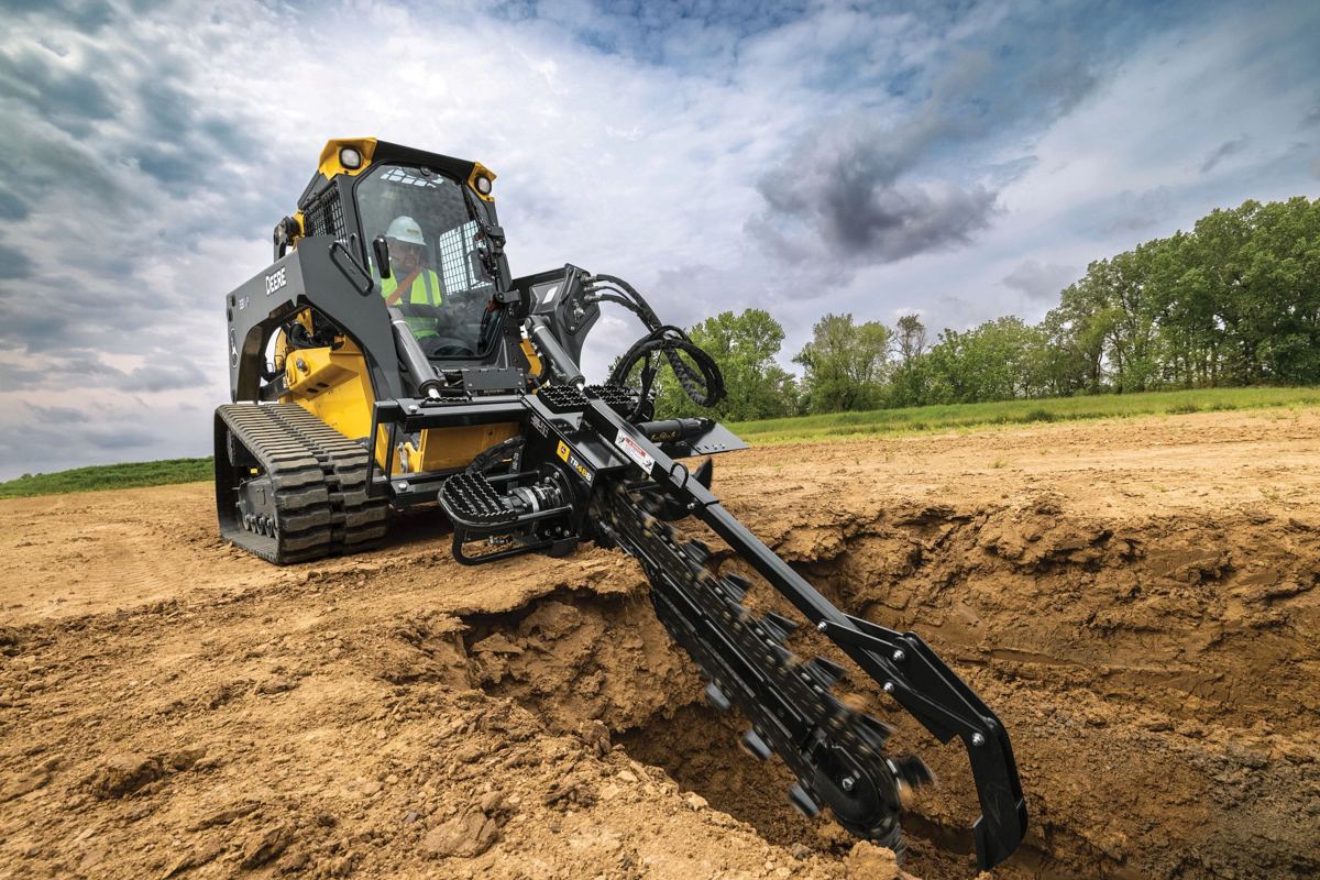 A John Deere compact track loader with a trencher attachment is actively digging a trench in brown soil under a cloudy sky