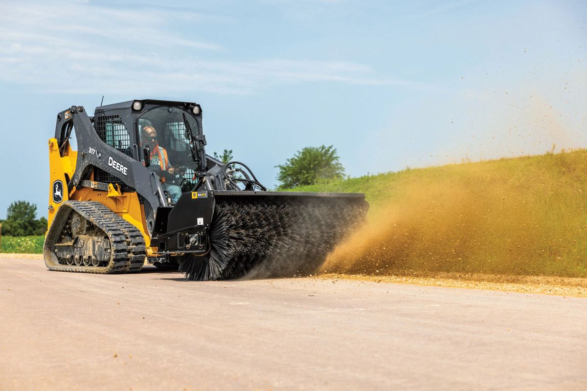 A John Deere compact track loader with an angle broom is actively sweeping a dirt road, kicking up a cloud of dust