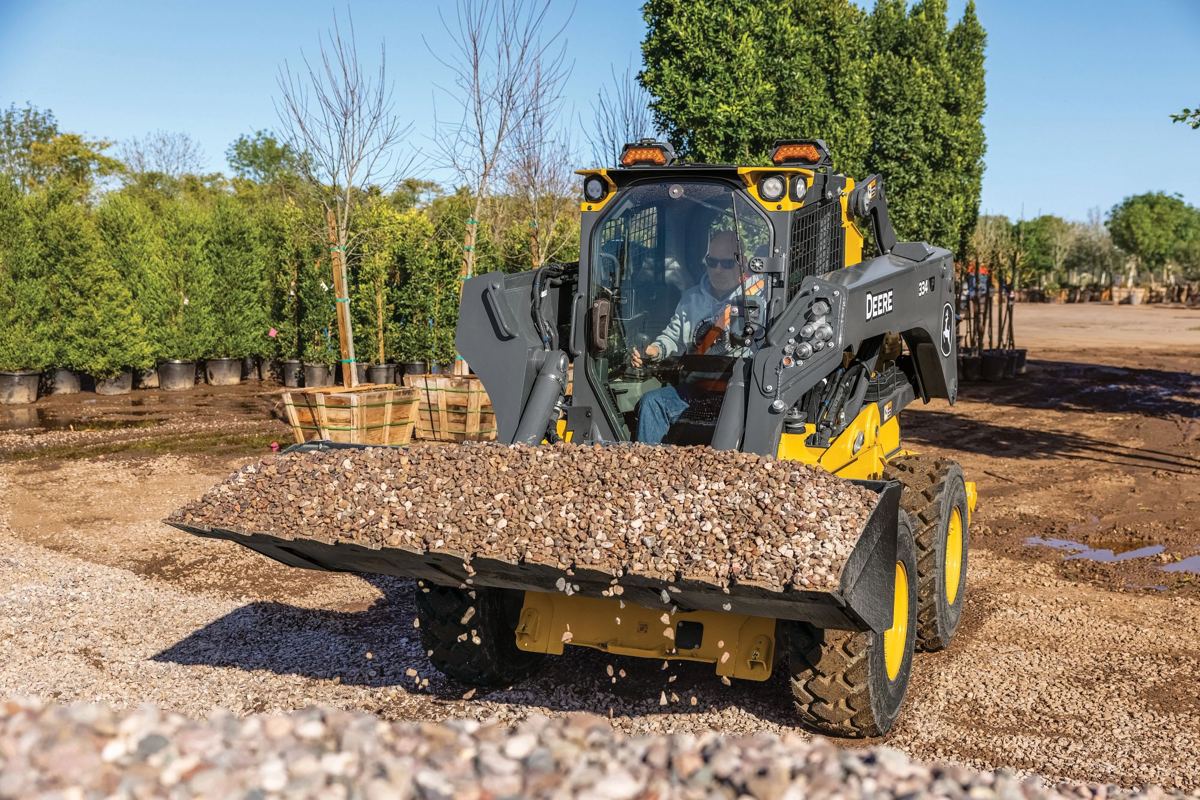A John Deere compact track loader with a bucket full of gravel, in use outdoors at a landscaping or nursery site