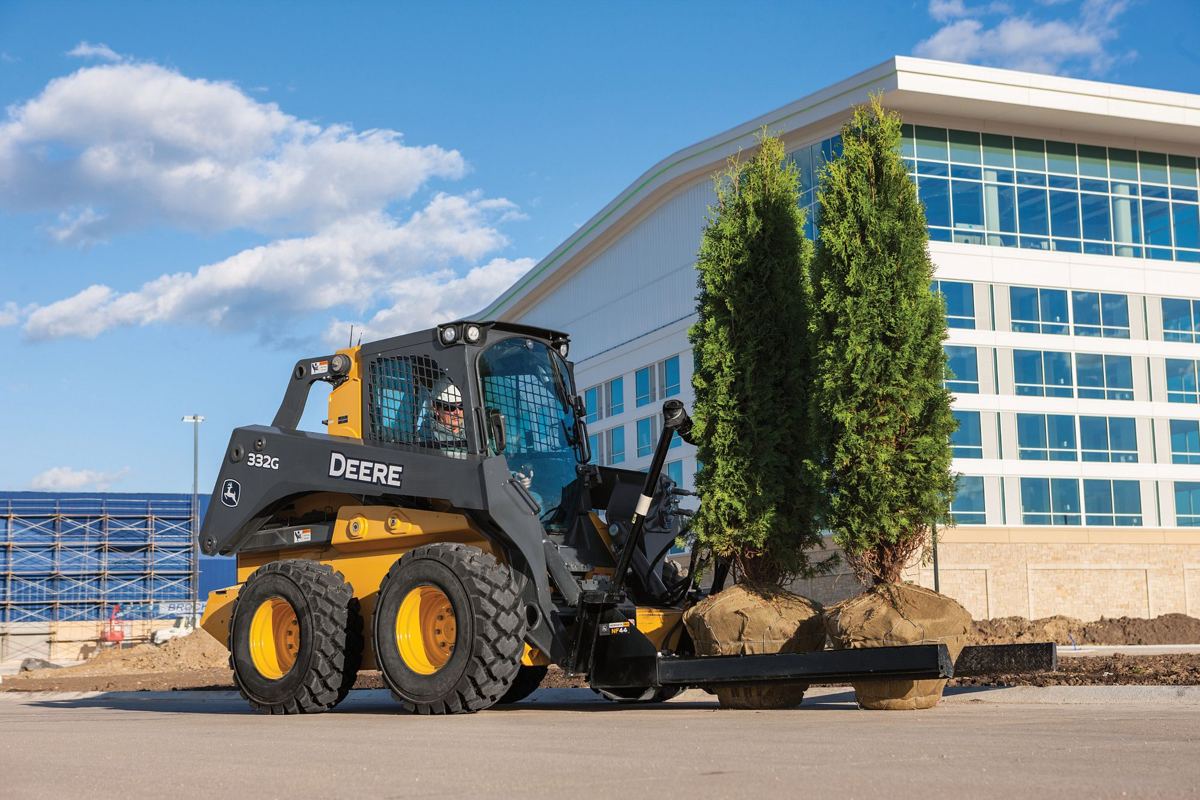 A John Deere skid steer loader with nursery forks is carrying two balled trees on a paved surface in front of a modern building