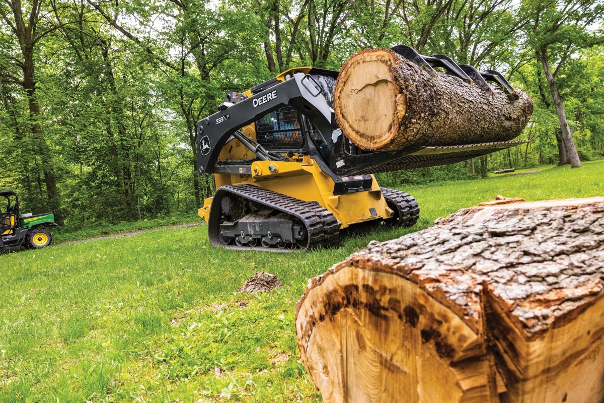A John Deere track loader with a grapple attachment is actively lifting and moving a large log in a wooded, grassy area, demonstrating clearing debris