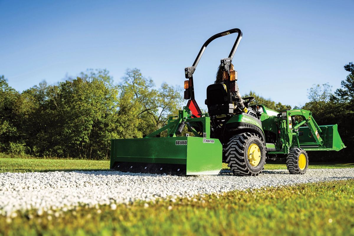 Person operating a green John Deere tractor with a Frontier BB5060 box blade and front loader on a gravel path