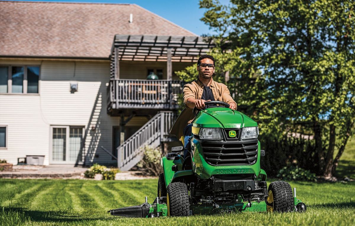 A man operates a John Deere X300 series lawn tractor, mowing a residential lawn with visible stripes