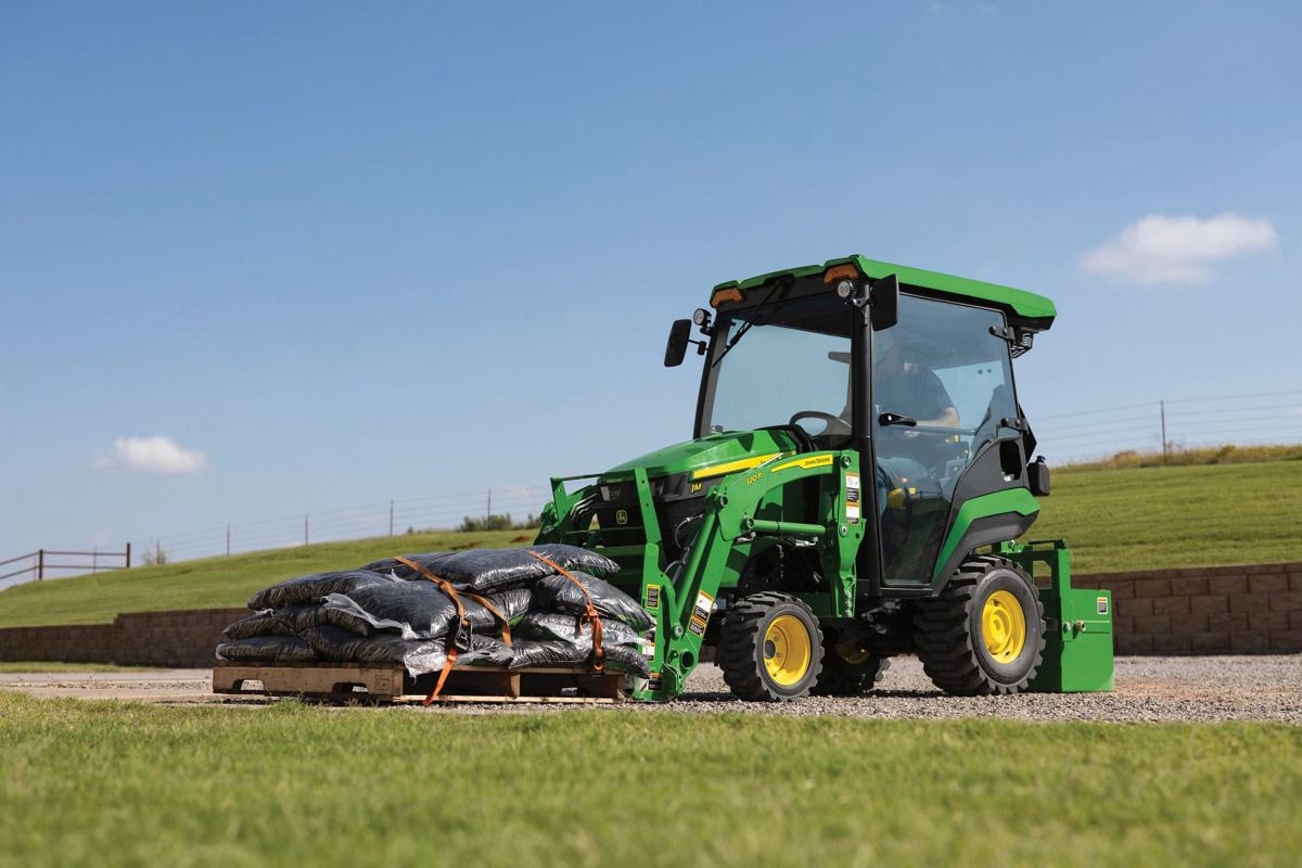 A John Deere compact tractor uses a front-end loader with pallet forks to move a pallet of bags
