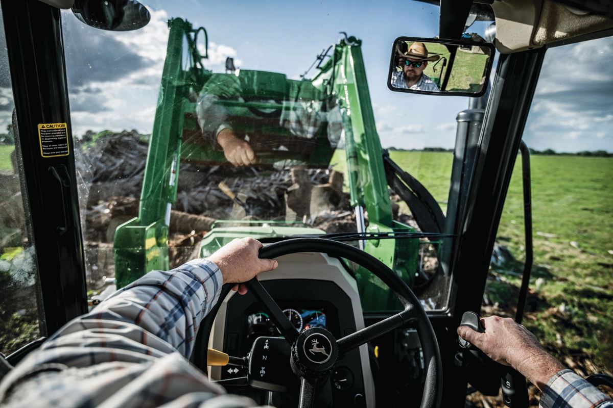 Man driving a John Deere tractor, operating a front loader moving debris, with the driver reflected in the rearview mirror