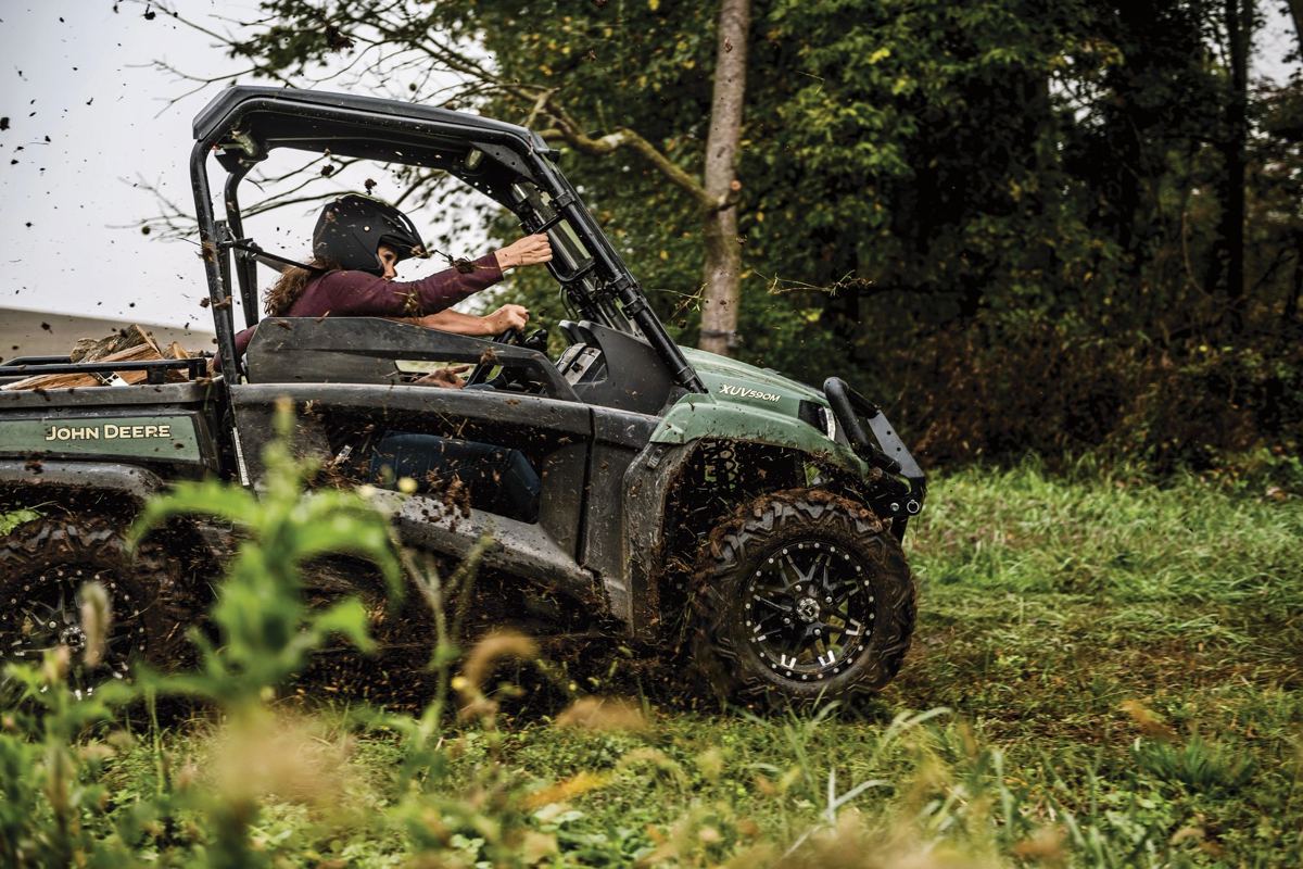 Person in a helmet driving a muddy green John Deere XUV590M utility vehicle through a grassy, wooded area