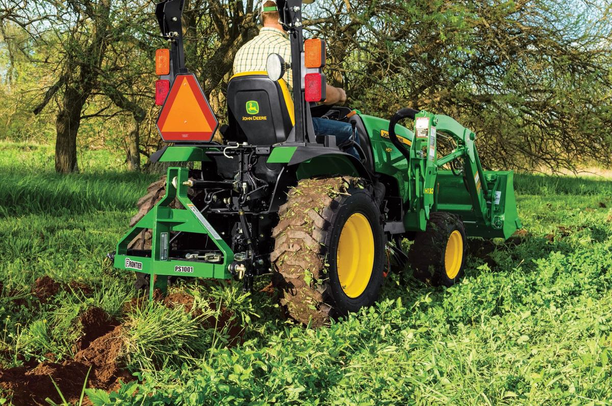 A John Deere compact utility tractor with a Frontier PS10 subsoiler working in a field with green vegetation