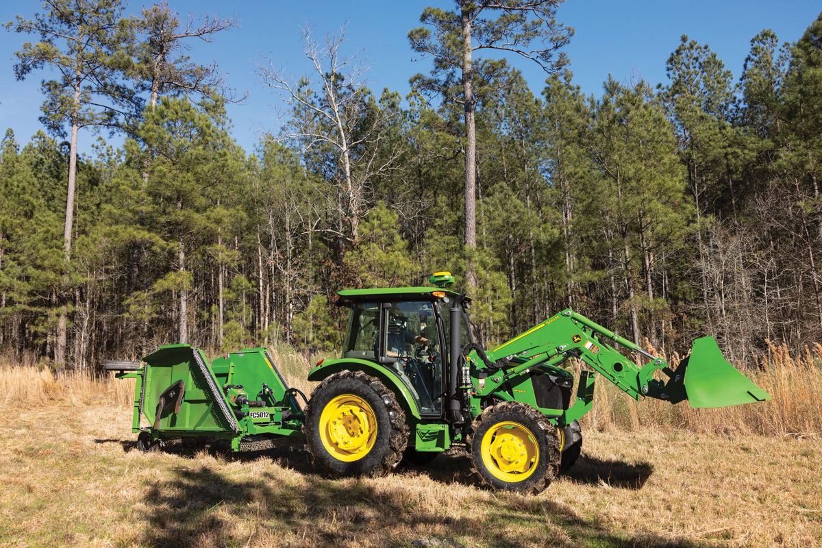 John Deere tractor with front loader and green FC5012 implement in a grassy field by pine trees