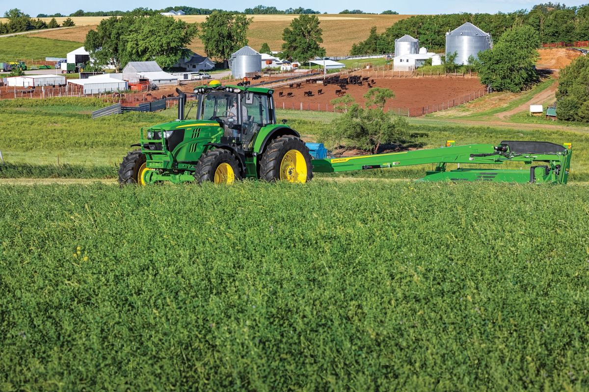 A green John Deere tractor pulls a green hay conditioner through a lush field with a farm and silos in the background