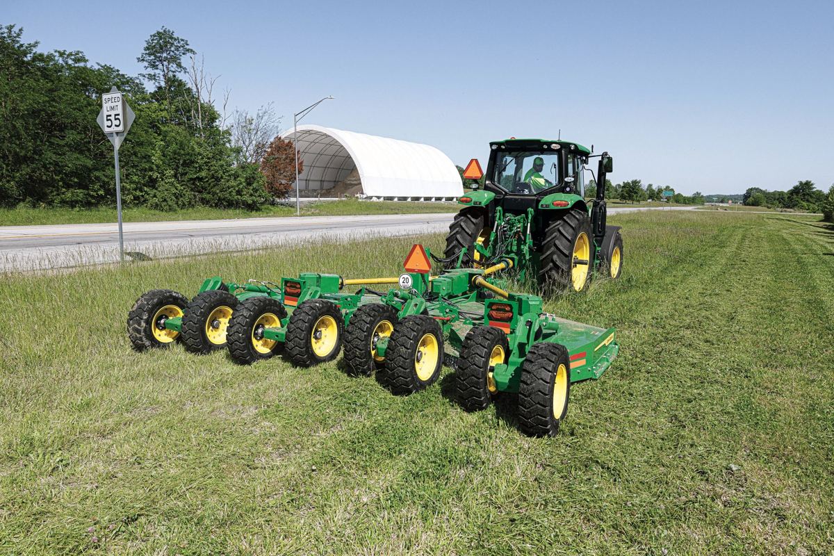 John Deere tractor pulling a wide rotary mower cutting grass on a roadside next to a highway