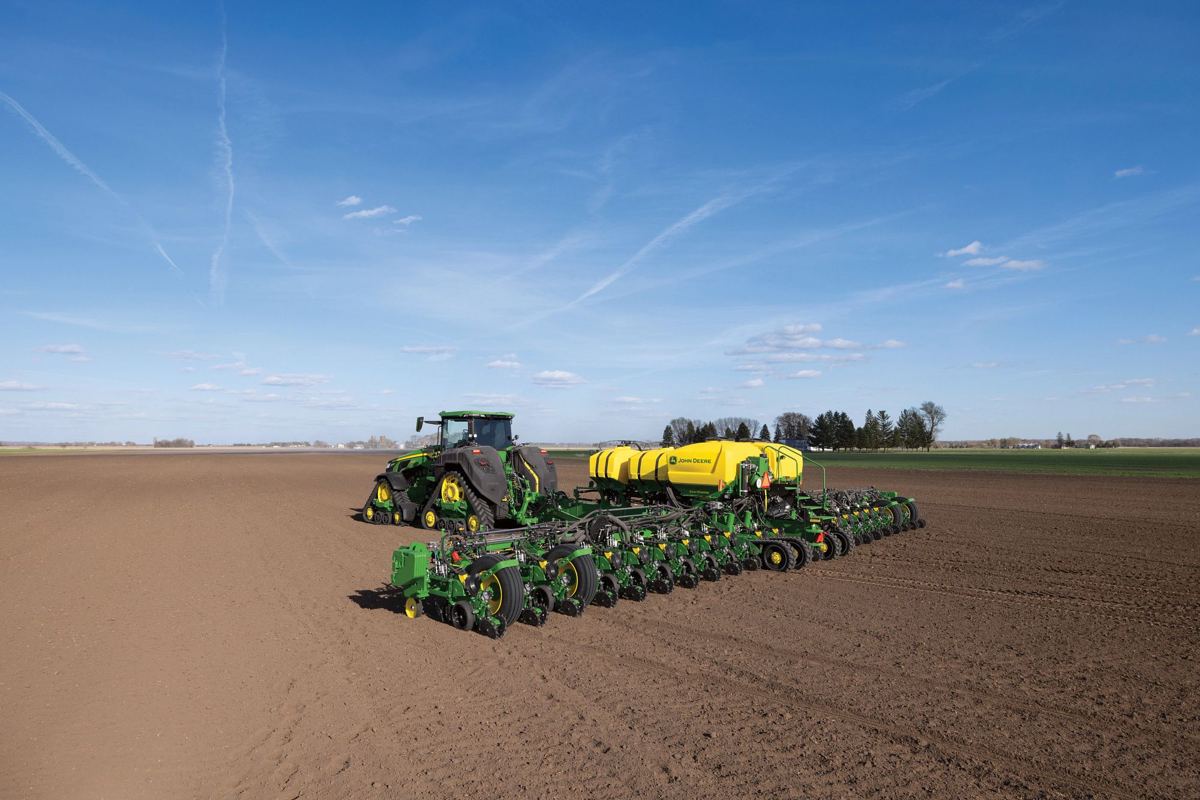 A John Deere tracked tractor pulls a large John Deere central-fill planter across a bare field under a blue sky, actively planting