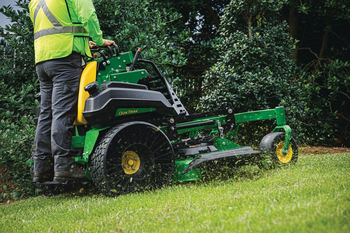 Person in safety vest operating a John Deere QuikTrak stand-on mower cutting grass