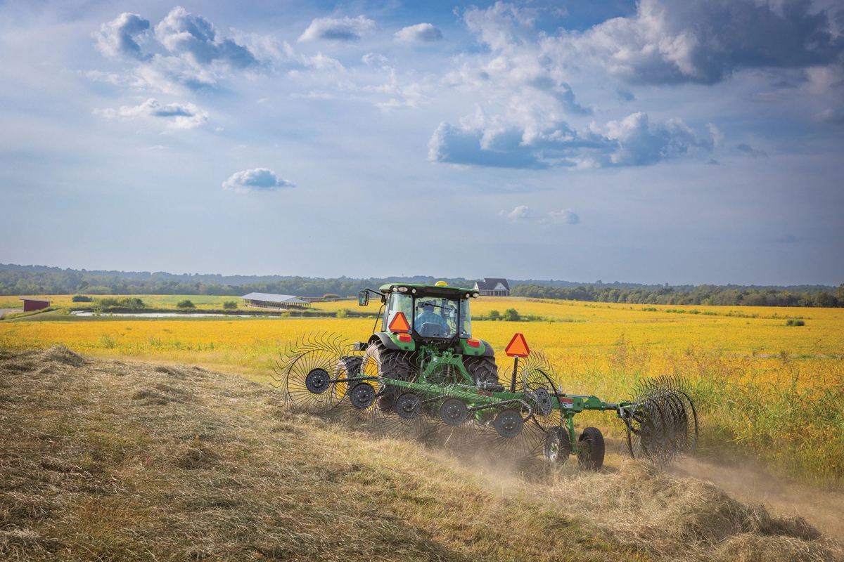 John Deere tractor with hay rake working in a field with yellow crops and buildings in the distance