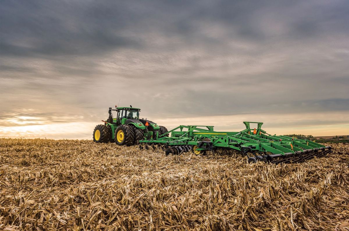 Green John Deere tractor pulling a large green tillage implement in a field of dry corn stalks under a cloudy sky