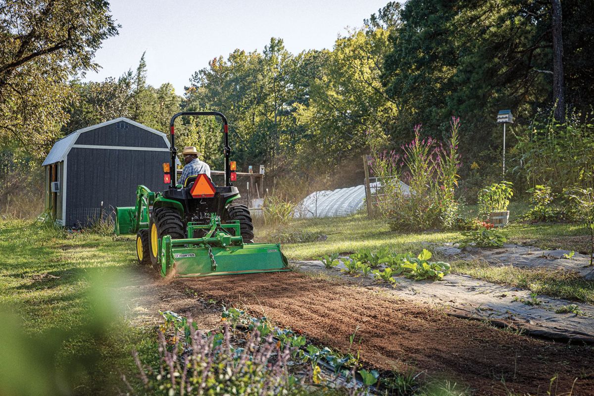 Man on John Deere tractor with Frontier tiller cultivating garden soil near a shed and greenhouse
