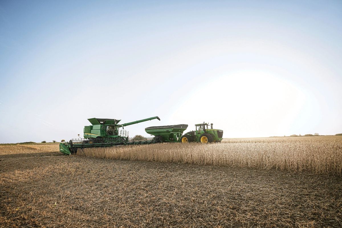 Green John Deere combine harvesting a field of crops, unloading into a grain cart pulled by a tractor